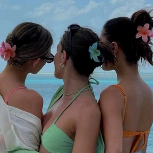 Three gradient frangipani flower hair claws worn by models against a blue ocean backdrop