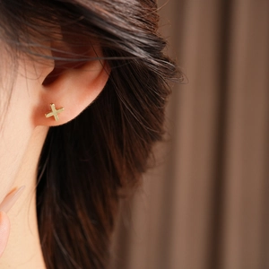 Close-up of a minimalist golden cross stud earring worn by a model in soft lighting