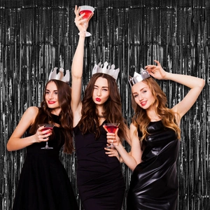 Three happy women holding cocktails in front of a shimmering black fringe curtain backdrop