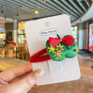 Détail d'une pince à cheveux pomme de Noël verte avec nœud rouge sur emballage