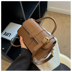 Brown leather handbag with gold buckle and top handle shown next to a book on a shelf