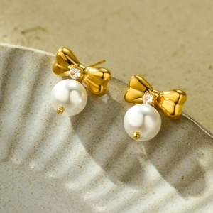 Elegant gold bow earrings with clear zircon and white pearls on ceramic tray