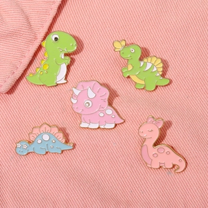 Five colorful cartoon enamel dinosaur brooches displayed on a pink denim background