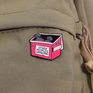 Broche en émail rose forme poubelle avec texte affichée sur un sac à dos kaki