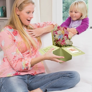 Woman and girl opening a green 3D pop-up flower bouquet birthday card in a warm living room