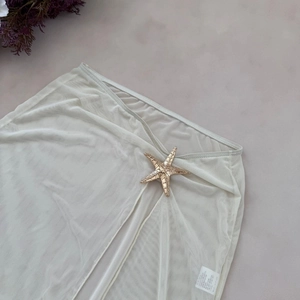 Detail of creamy mesh beach skirt featuring a golden starfish buckle on a textured background
