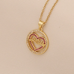 Golden heart necklace with MOM text and red crystals on beige fabric background