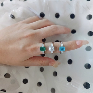 Close-up of three emerald-cut moissanite rings in green, clear, and aqua blue on a hand