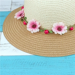 Close-up of beige straw sun hat with pink daisy wreath and green leaves on wood
