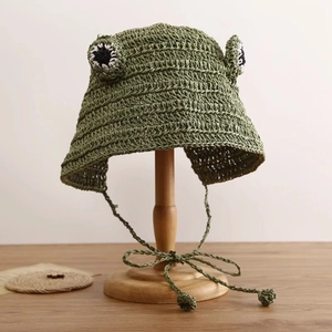 Close-up of green crochet frog hat for kids with eye details and chin straps on wood stand
