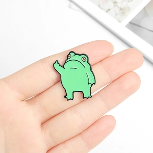 Funny green frog enamel pin making a middle finger gesture held in hand on white background