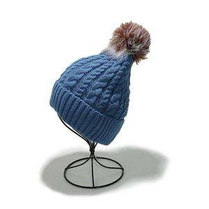 Close-up of blue cable-knit beanie with a brown faux fur pompom on a display stand