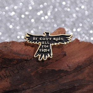 Macro detail of black crow enamel pin with It Can't Rain All The Time gold engraving