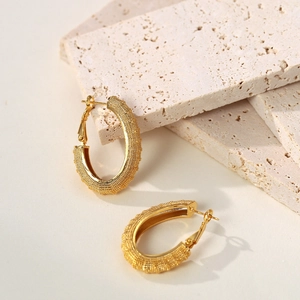 Elegant gold oval hoop earrings with textured grainy finish on minimalist stone background