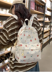 Rear view of a white canvas backpack with strawberry prints and a white fuzzy pompom charm