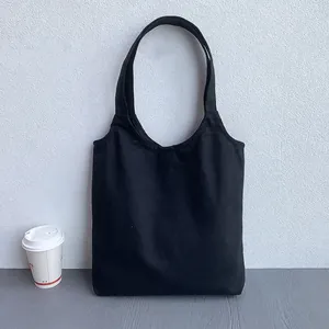 Minimalist black canvas tote bag with wide straps shown in a lifestyle coffee shop setting