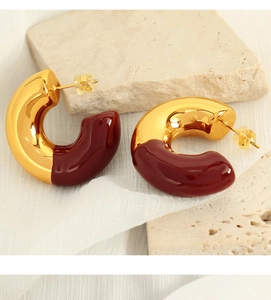Detail of C-shaped gold metal earrings with burgundy enamel on beige stone background