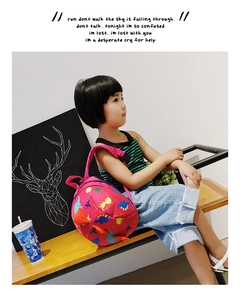 Pink cartoon dinosaur toddler backpack worn by a little girl sitting on a wooden bench