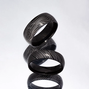 Macro shot of 6mm and 8mm black Damascus steel wedding bands on a grey reflective surface