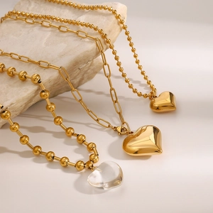 Three gold heart pendant necklaces featuring paperclip and bead chains on stone