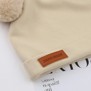 Close-up of beige knitted baby hat showing fluffy pom-pom and brown handmade leather label