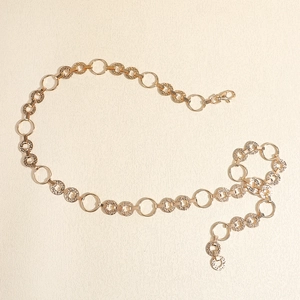 Dettaglio di una cintura a catena dorata con anelli circolari testurizzati e chiusura a moschettone