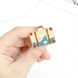 Beige enamel travel suitcase brooch with blue wave pattern and brown leather straps