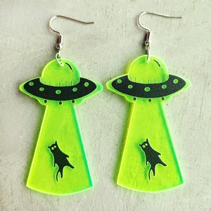 Detail of neon green acrylic UFO and cat earrings laying on a light textured surface
