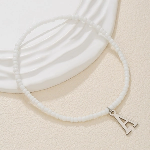 Close-up of a white seed bead anklet featuring a silver letter A pendant on beige surface