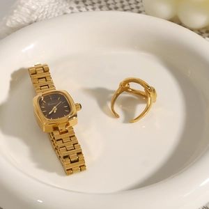Detail shot of a square gold watch and a twisted open ring on a white plate