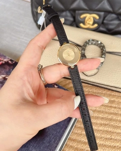 Close-up of a minimalist gold watch with black leather strap held by a model in soft light