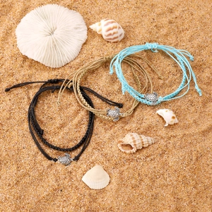 Three handmade woven turtle anklets in black, beige, and blue on a sandy beach