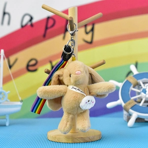 Detail view of a cute beige plush bunny keychain with a rainbow strap on a stand.