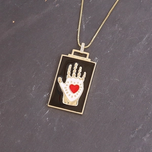 Macro detail of gold square pendant with black enamel, white hand and red heart motif