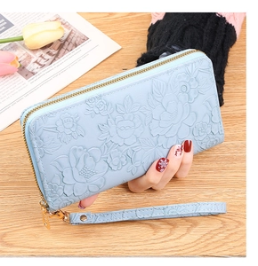 Blue long floral embossed wallet with gold zipper and wristlet held over a wooden desk