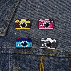 Dettaglio di spillette in smalto a forma di fotocamera retrò in quattro colori su denim