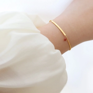 Close-up of gold beaded bracelet with rectangular bar and red teardrop charm on model