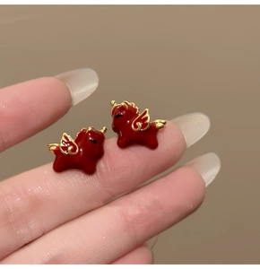 Macro view of small red enamel winged horse earrings held between fingers for scale