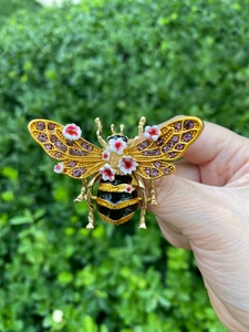 Detailed view of a gold bee brooch with pink rhinestones held against a garden background
