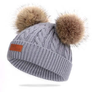 Détail du bonnet gris en maille torsadée avec deux pompons en fourrure et étiquette