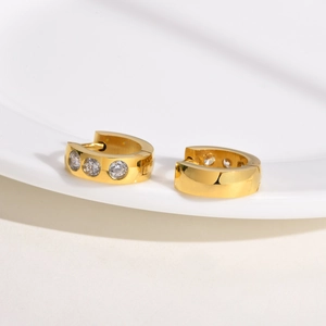 Close-up of small gold huggie hoop earrings with three inlaid white zircons on ceramic