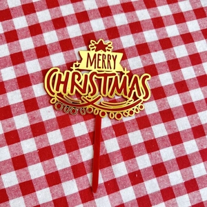 Macro shot of red and gold acrylic Merry Christmas cake topper on red plaid fabric
