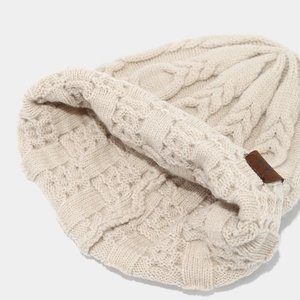 Détail du bonnet en maille crème avec torsades épaisses et patch logo en cuir marron