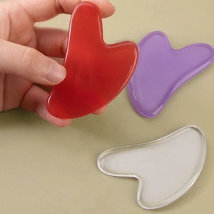 Three heart-shaped resin facial scrapers in red, purple, and transparent held by hand