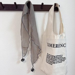 Detail of grey triangle scarf with pom-poms hanging on a hook next to a canvas tote bag