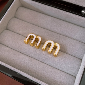 Macro view of gold-plated letter M earrings with white enamel inlay on grey velvet