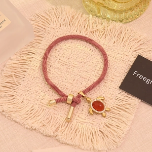 Detail eines rosa gewebten Armbands mit goldenem Schildkrötenanhänger und rotem Stein