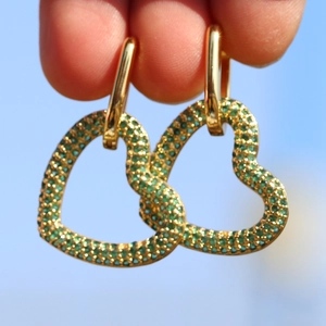 Orecchini a cuore placcati oro 18K con zirconi verdi tenuti contro lo sfondo del cielo blu