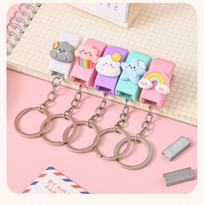 Flat lay of five mini cartoon stapler keychains in pastel colors on a pink notebook