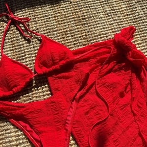 Flat lay of red three-piece bikini set with matching mesh cover-up skirt on a straw mat